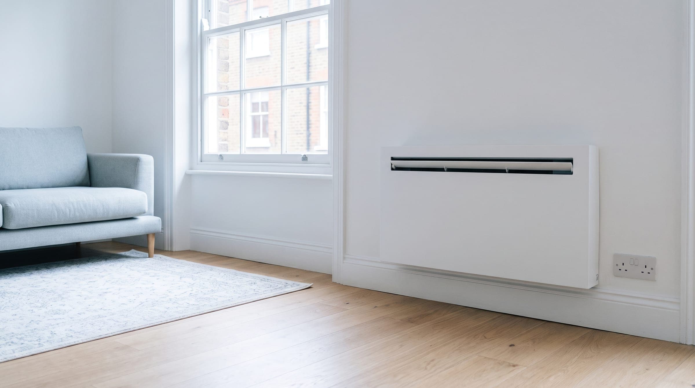 White through-wall air conditioning unit fitted low on the wall of a bright London flat living room, with no outdoor unit