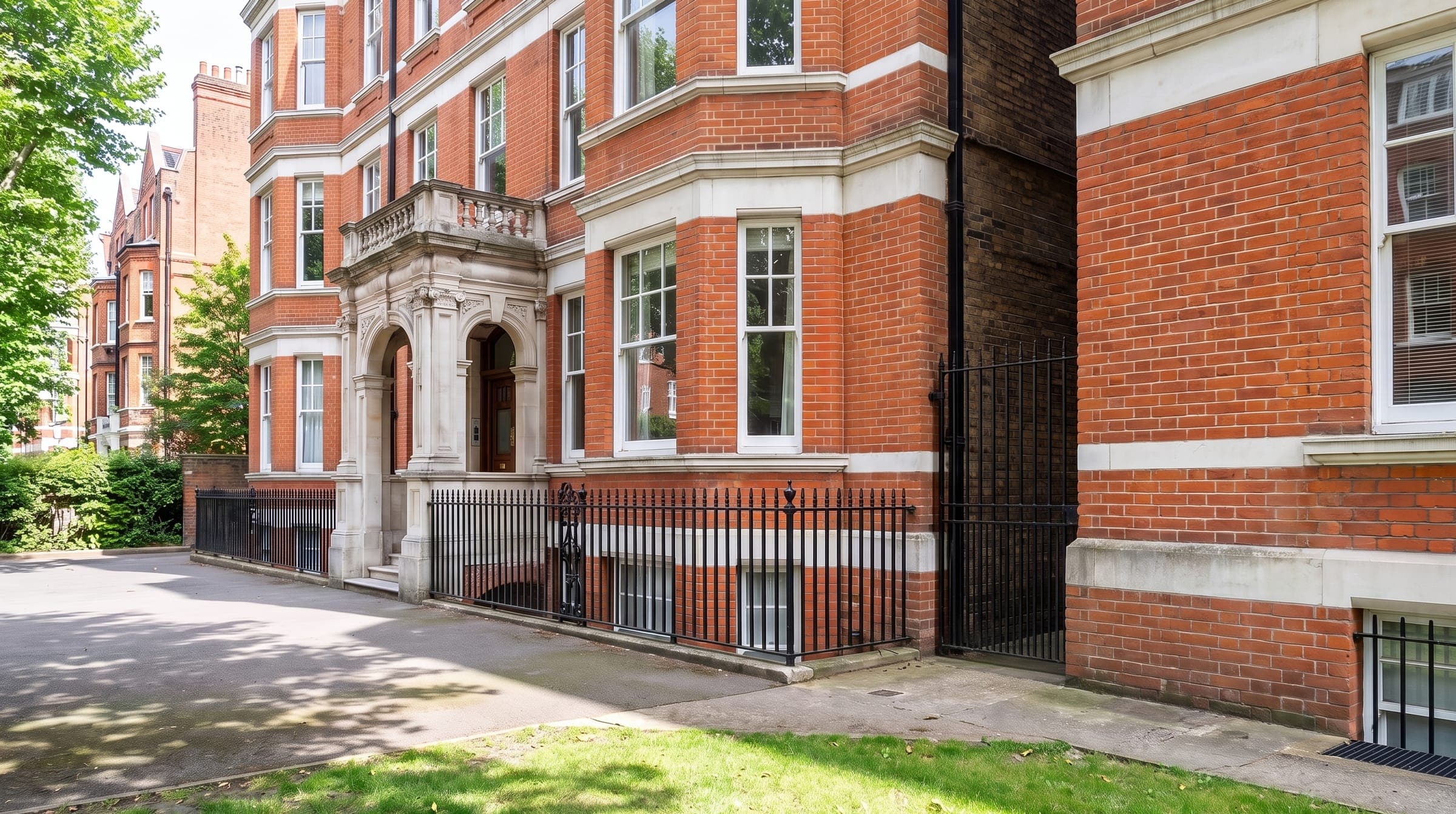 Red-brick Victorian mansion block with white stone bands, bay windows, black railings and a gated side passage on a leafy street