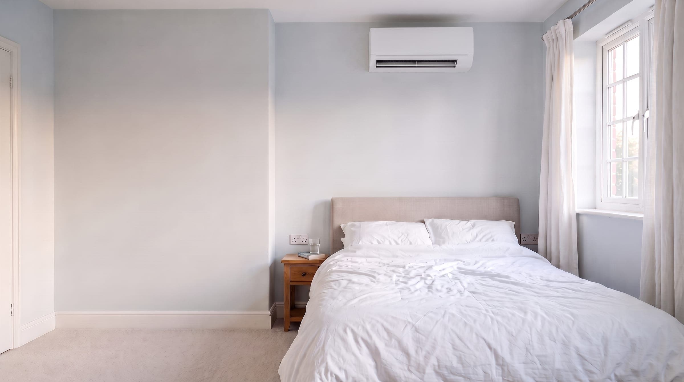 Calm, cool UK bedroom on a bright summer evening, light curtains, crisp white bedding and a white wall unit above the bed with its flap open
