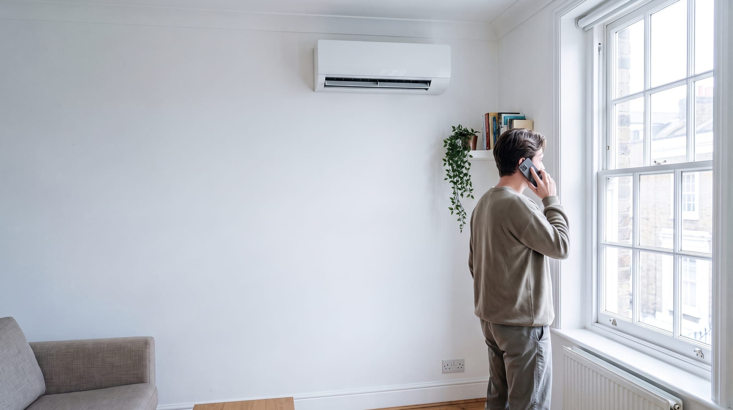 Young tenant in a bright rented London flat talking on the phone by the window, with a white air conditioning wall unit high on the wall