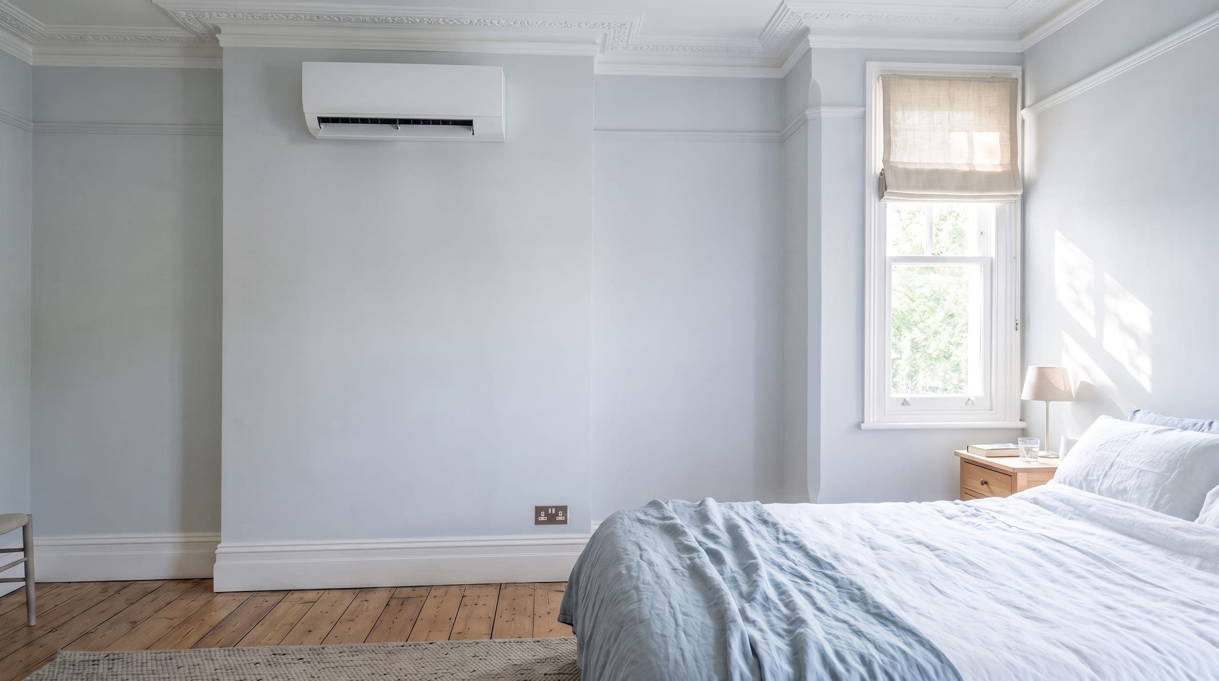 Slim white wall-mounted air conditioning unit high on the wall of a bright Victorian bedroom, its flap open and running